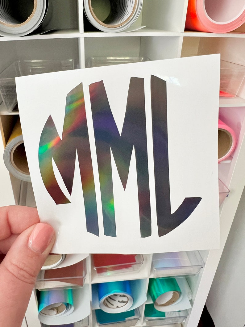 May include: A vinyl decal featuring the letters MML in a stylized, bold font. The letters have a holographic finish that reflects a spectrum of colors including green, yellow, and red against a dark, metallic base on a white backing sheet.