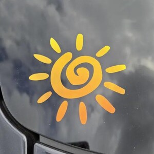 Puede incluir: Una calcoman&iacute;a de sol amarilla con un dise&ntilde;o de espiral central rodeado por doce rayos radiantes de longitudes variables. El gr&aacute;fico est&aacute; aplicado sobre una superficie oscura y reflectante, creando un contraste con el color amarillo brillante.