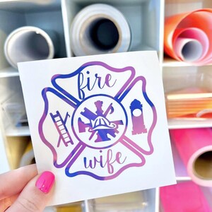 May include: A square white decal featuring a purple and blue gradient Maltese cross emblem. The design includes a central firefighter helmet, a ladder on the left, a tower on the right, and the text "fire" above and "wife" below in script.