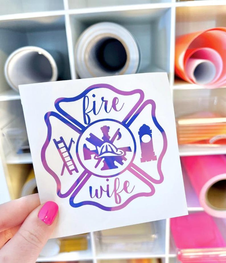 May include: A square white decal featuring a purple and blue gradient Maltese cross emblem. The design includes a central firefighter helmet, a ladder on the left, a tower on the right, and the text "fire" above and "wife" below in script.