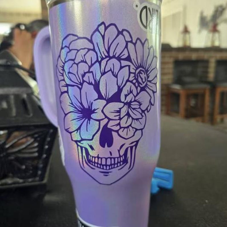 Puede incluir: Un vaso lavanda con acabado hologr&aacute;fico, con un dise&ntilde;o de calavera floral en morado. El vaso tiene un asa y una pegatina en el lateral. El dise&ntilde;o incluye varias formas de flores y una calavera detallada.