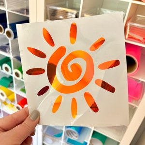 Puede incluir: Una hoja cuadrada de papel protector blanco con una calcoman&iacute;a de sol con acabado naranja met&aacute;lico. El dise&ntilde;o del sol consta de un n&uacute;cleo central en espiral rodeado por doce rayos ovalados alargados y distintos en c&iacute;rculo.