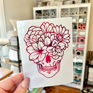 Puede incluir: Una pegatina de calavera floral roja y rosa. La calavera est&aacute; representada con un arreglo floral detallado en lugar de cabello. La pegatina se sostiene frente a un fondo blanco.