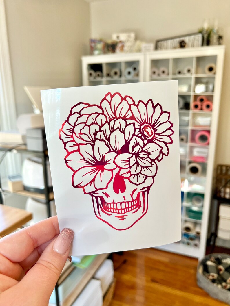 Puede incluir: Una pegatina de calavera floral roja y rosa. La calavera est&aacute; representada con un arreglo floral detallado en lugar de cabello. La pegatina se sostiene frente a un fondo blanco.