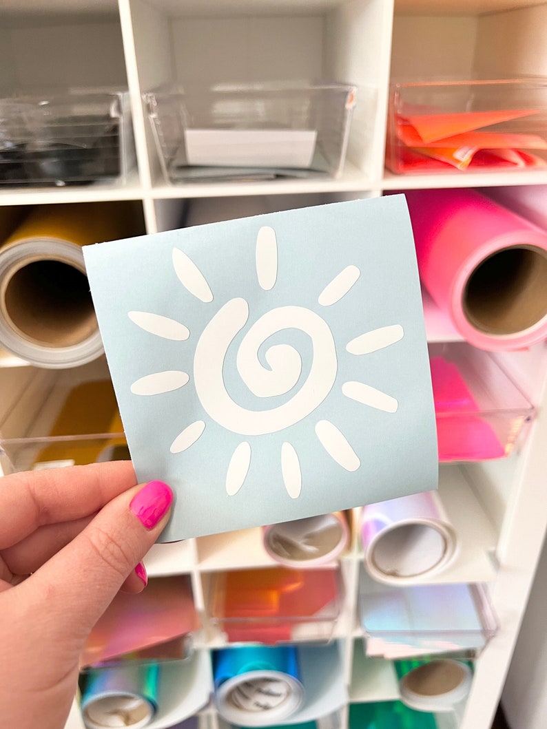 Puede incluir: Una calcoman&iacute;a de vinilo cuadrada de color azul claro con un gr&aacute;fico de sol blanco que presenta un centro en espiral y rayos ovalados. La calcoman&iacute;a se sostiene frente a un organizador de suministros con rollos de vinilo de colores.
