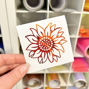 May include: A square white decal featuring a stylized sunflower graphic with a metallic orange and red gradient finish. The sunflower design consists of pointed petals surrounding a central circular core filled with small white dots.