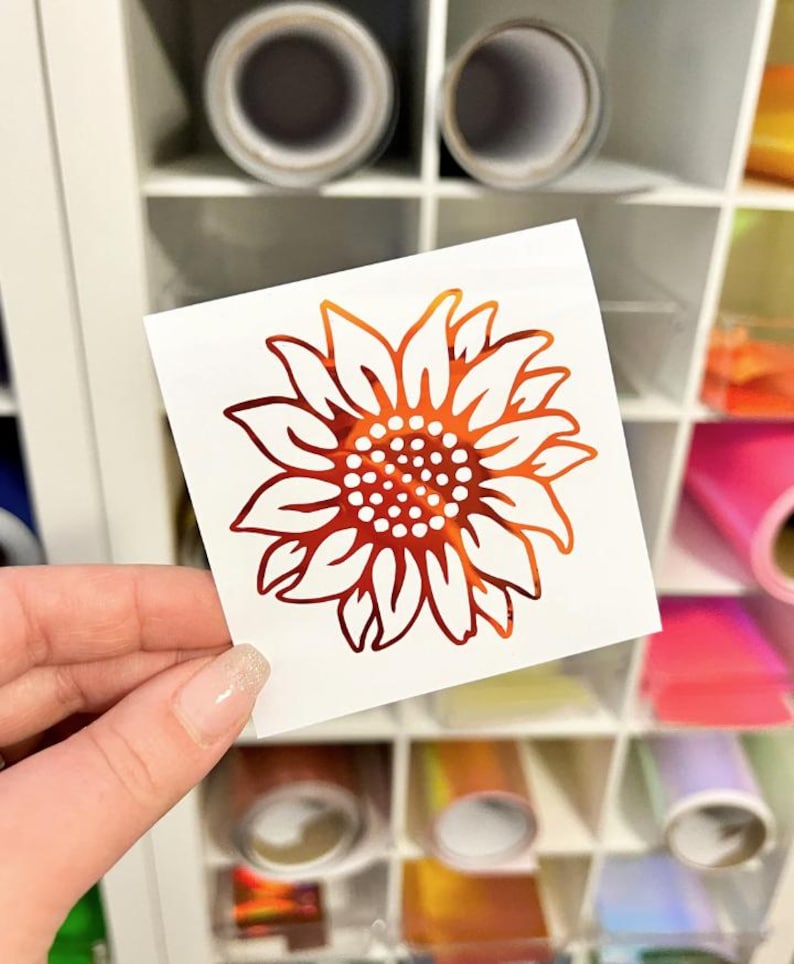 May include: A square white decal featuring a stylized sunflower graphic with a metallic orange and red gradient finish. The sunflower design consists of pointed petals surrounding a central circular core filled with small white dots.