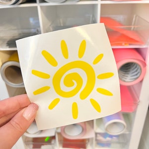 Puede incluir: Una calcoman&iacute;a cuadrada blanca que presenta un gr&aacute;fico de sol amarillo brillante con una espiral central y doce rayos radiantes. El dise&ntilde;o del sol es simple y audaz, impreso en una hoja de respaldo lisa y brillante sostenida frente a un mueble.