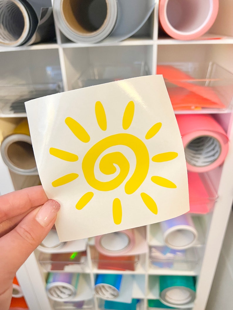 Puede incluir: Una calcoman&iacute;a cuadrada blanca que presenta un gr&aacute;fico de sol amarillo brillante con una espiral central y doce rayos radiantes. El dise&ntilde;o del sol es simple y audaz, impreso en una hoja de respaldo lisa y brillante sostenida frente a un mueble.