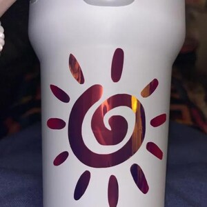 Puede incluir: Un vaso blanco con un gr&aacute;fico central de sol con un centro en espiral. El dise&ntilde;o del sol est&aacute; realizado en un acabado met&aacute;lico iridiscente que cambia entre tonos rojo oscuro, morado y naranja, contrastando con la superficie blanca mate.