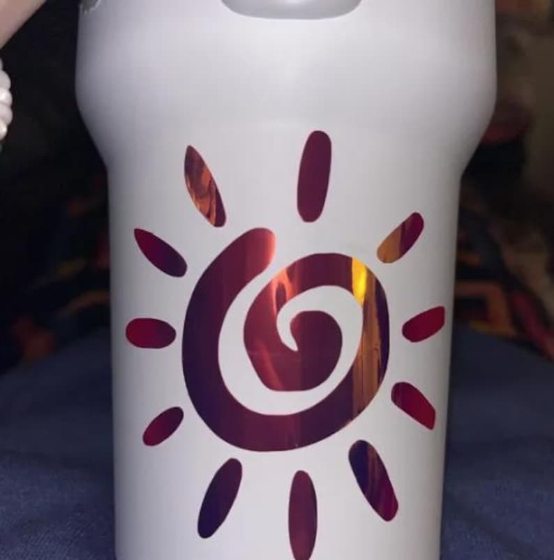 Puede incluir: Un vaso blanco con un gr&aacute;fico central de sol con un centro en espiral. El dise&ntilde;o del sol est&aacute; realizado en un acabado met&aacute;lico iridiscente que cambia entre tonos rojo oscuro, morado y naranja, contrastando con la superficie blanca mate.