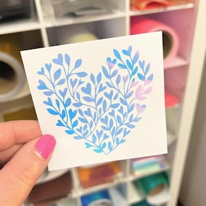 May include: A square white sticker featuring a heart-shaped design composed of blue and light purple botanical branches with small leaves. The sticker displays a gradient color effect transitioning from deep blue to soft purple across the foliage.