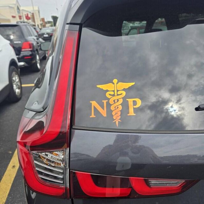 May include: A gold-colored decal applied to the rear window of a dark gray vehicle. The decal features a caduceus symbol centered between the capital letters N and P, all rendered in a bold, sans-serif font with a metallic yellow finish.