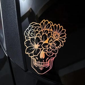 Puede incluir: Una pegatina de calavera dorada y naranja con elementos florales. La pegatina presenta un dise&ntilde;o de calavera con flores y otros elementos florales. La pegatina est&aacute; adherida a una superficie negra.