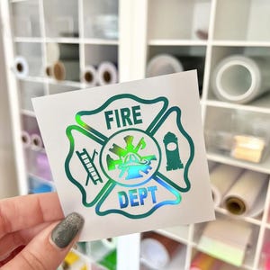 May include: A square white decal featuring a holographic, iridescent fire department emblem. The design includes a central fire helmet, a ladder, and a fire hydrant, with the text FIRE DEPT printed in bold, capital letters within the cross shape.