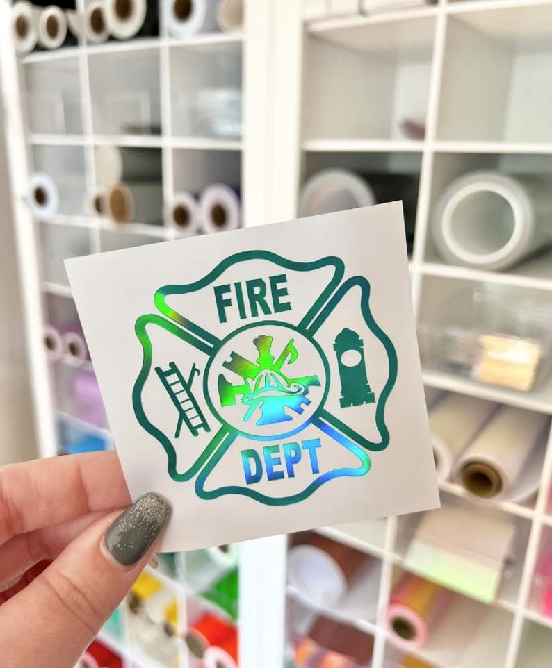 May include: A square white decal featuring a holographic, iridescent fire department emblem. The design includes a central fire helmet, a ladder, and a fire hydrant, with the text FIRE DEPT printed in bold, capital letters within the cross shape.