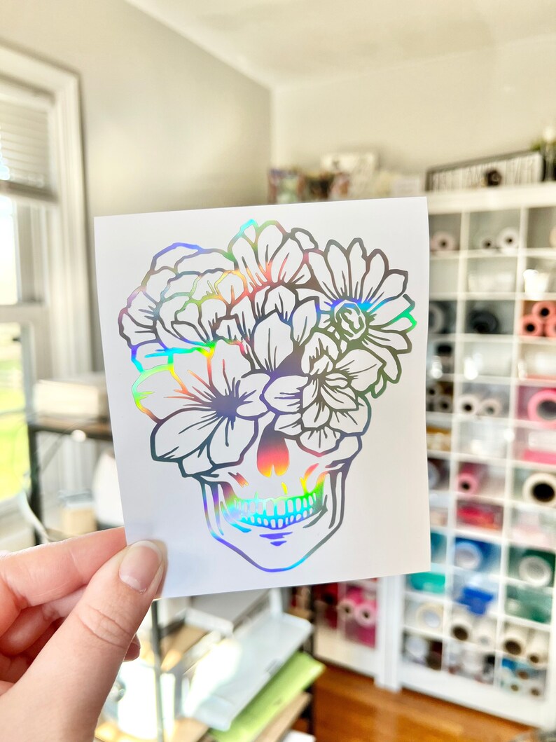 Puede incluir: Una pegatina hologr&aacute;fica con un dise&ntilde;o de calavera y elementos florales. La calavera est&aacute; adornada con varias flores. La pegatina tiene un acabado iridiscente reflectante, creando un efecto arco&iacute;ris.