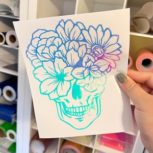 Puede incluir: Una pegatina blanca con un dise&ntilde;o de calavera con una corona floral. Las flores y la calavera tienen un degradado de azul, turquesa y morado. La pegatina se sostiene frente a una estanter&iacute;a blanca.