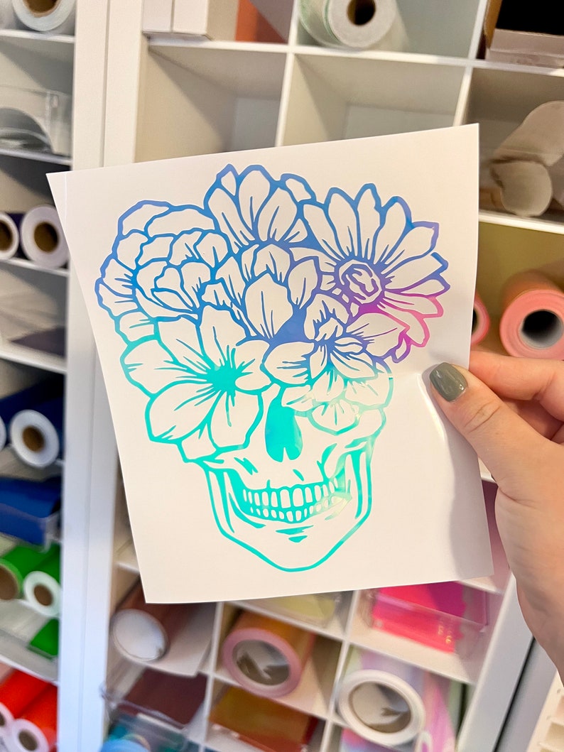 Puede incluir: Una pegatina blanca con un dise&ntilde;o de calavera con una corona floral. Las flores y la calavera tienen un degradado de azul, turquesa y morado. La pegatina se sostiene frente a una estanter&iacute;a blanca.
