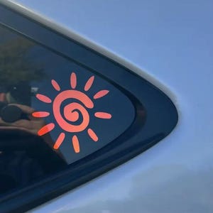 Puede incluir: Un adhesivo de sol de color naranja brillante con un dise&ntilde;o de espiral central rodeado por doce rayos ovalados radiantes, aplicado sobre el cristal tintado oscuro de la ventana de un veh&iacute;culo. La pegatina tiene un acabado liso y vibrante.