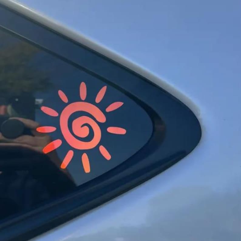 Puede incluir: Un adhesivo de sol de color naranja brillante con un dise&ntilde;o de espiral central rodeado por doce rayos ovalados radiantes, aplicado sobre el cristal tintado oscuro de la ventana de un veh&iacute;culo. La pegatina tiene un acabado liso y vibrante.