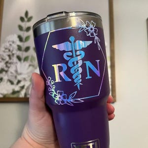 May include: A purple stainless steel tumbler with a clear lid and a silver YETI logo at the base. The front features a holographic decal of a medical caduceus symbol between the letters R and N, enclosed in a hexagon frame with floral accents.