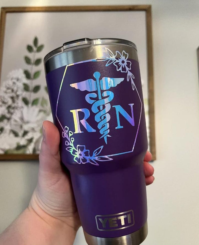 May include: A purple stainless steel tumbler with a clear lid and a silver YETI logo at the base. The front features a holographic decal of a medical caduceus symbol between the letters R and N, enclosed in a hexagon frame with floral accents.