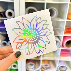 May include: A square white decal featuring a sunflower design with a holographic finish that reflects a spectrum of colors. The sunflower has a central cluster of small circles surrounded by pointed petals, all rendered in a reflective material.