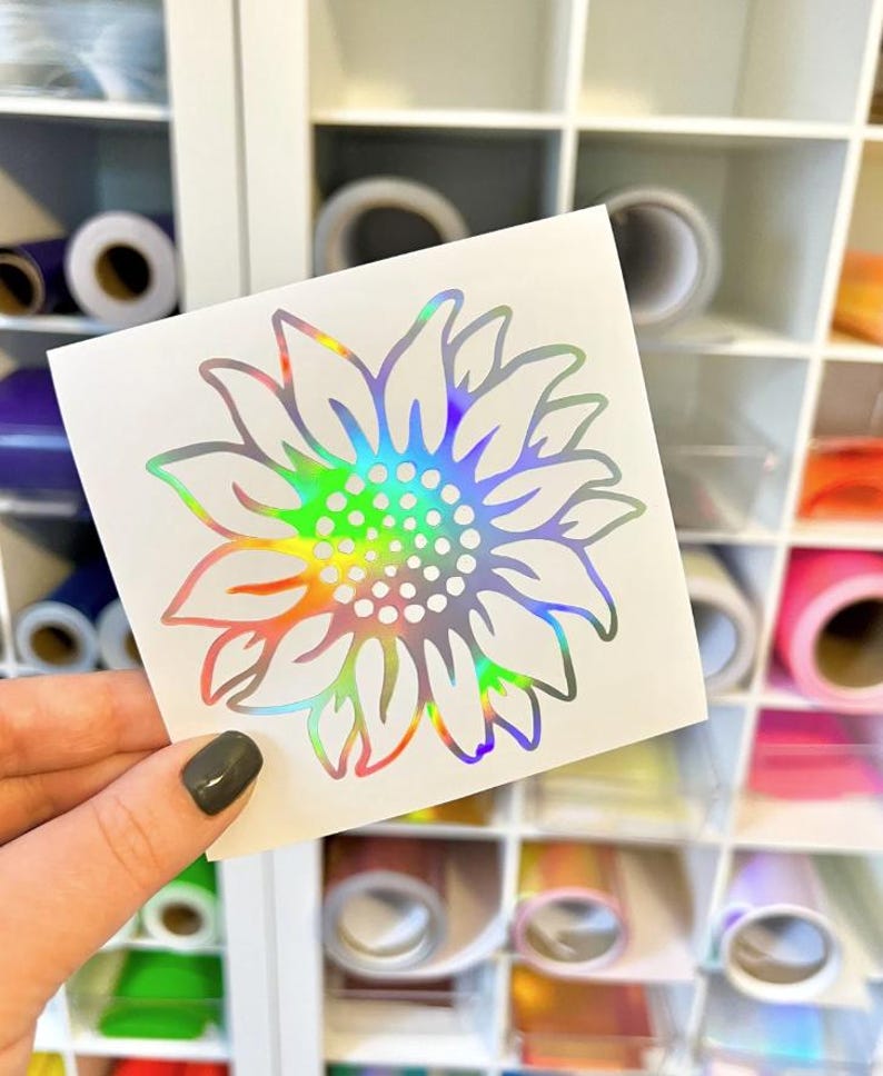 May include: A square white decal featuring a sunflower design with a holographic finish that reflects a spectrum of colors. The sunflower has a central cluster of small circles surrounded by pointed petals, all rendered in a reflective material.