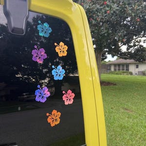 May include: A collection of seven hibiscus flower decals applied to a vehicle window. The stickers feature a variety of colors including iridescent blue, purple, orange, and pink, arranged in a scattered pattern across the dark glass surface.