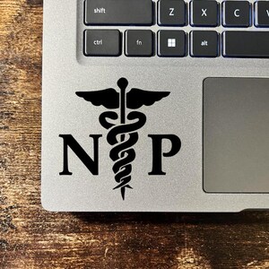 May include: A black vinyl decal applied to a silver laptop surface. The decal features the letters N and P flanking a central caduceus symbol, which consists of a winged staff entwined by two snakes. The design is rendered in a solid black color.