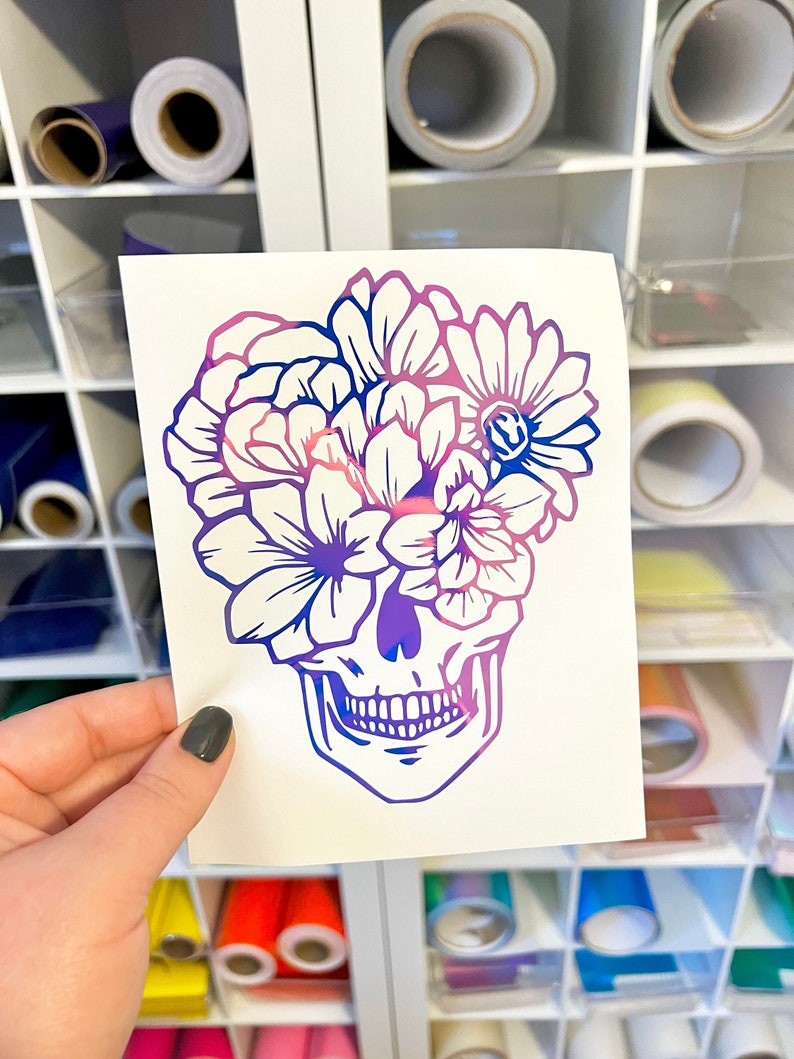 Puede incluir: Una pegatina blanca con un dise&ntilde;o de calavera con una corona floral. La calavera y las flores est&aacute;n en un degradado de p&uacute;rpura, rosa y azul. La pegatina se sostiene contra una estanter&iacute;a blanca con rollos de vinilo.
