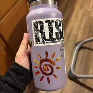 Puede incluir: Una botella de agua aislante de color morado claro con una pegatina rectangular que muestra el texto RISE, un n&uacute;mero de tel&eacute;fono y una direcci&oacute;n web. Debajo del texto hay una pegatina de sol naranja met&aacute;lico con un centro en espiral.