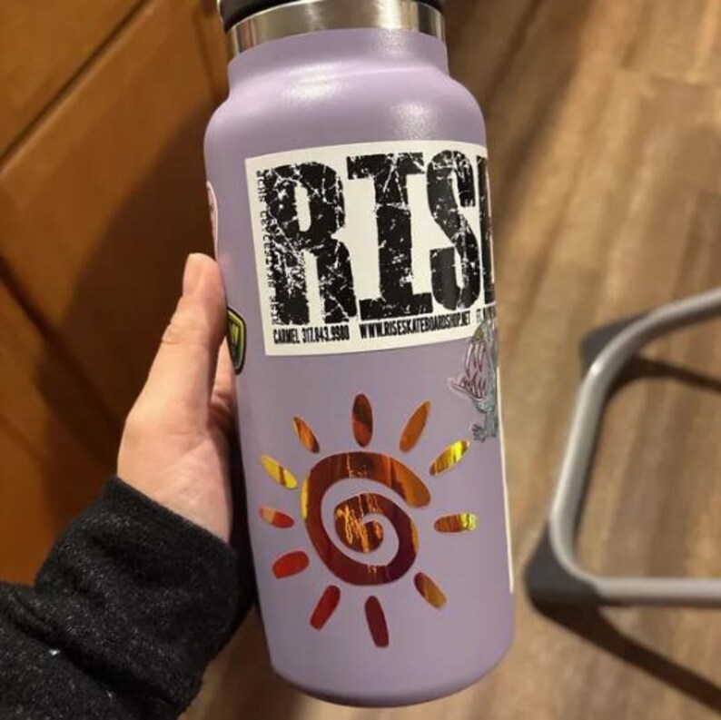Puede incluir: Una botella de agua aislante de color morado claro con una pegatina rectangular que muestra el texto RISE, un n&uacute;mero de tel&eacute;fono y una direcci&oacute;n web. Debajo del texto hay una pegatina de sol naranja met&aacute;lico con un centro en espiral.