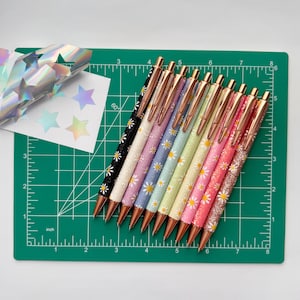 May include: A collection of ten ballpoint pens with daisy patterns in various colors, including black, white, pink, and blue. The pens have rose gold accents and are arranged on a green cutting mat with inch measurements.