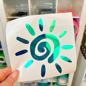 Puede incluir: Una hoja adhesiva blanca cuadrada con un dise&ntilde;o de sol troquelado. El sol tiene una espiral central y doce rayos circundantes, todo hecho de un material reflectante e iridiscente que cambia entre tonos de azul, verde y verde azulado.
