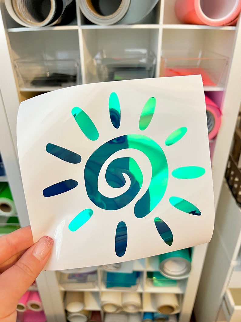 Puede incluir: Una hoja adhesiva blanca cuadrada con un dise&ntilde;o de sol troquelado. El sol tiene una espiral central y doce rayos circundantes, todo hecho de un material reflectante e iridiscente que cambia entre tonos de azul, verde y verde azulado.