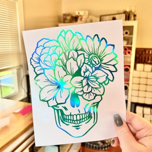 Puede incluir: Una pegatina hologr&aacute;fica con un dise&ntilde;o de calavera y elementos florales. La calavera est&aacute; delineada en verde y azul, con flores que florecen desde la parte superior. La pegatina se sostiene sobre un fondo brillante.