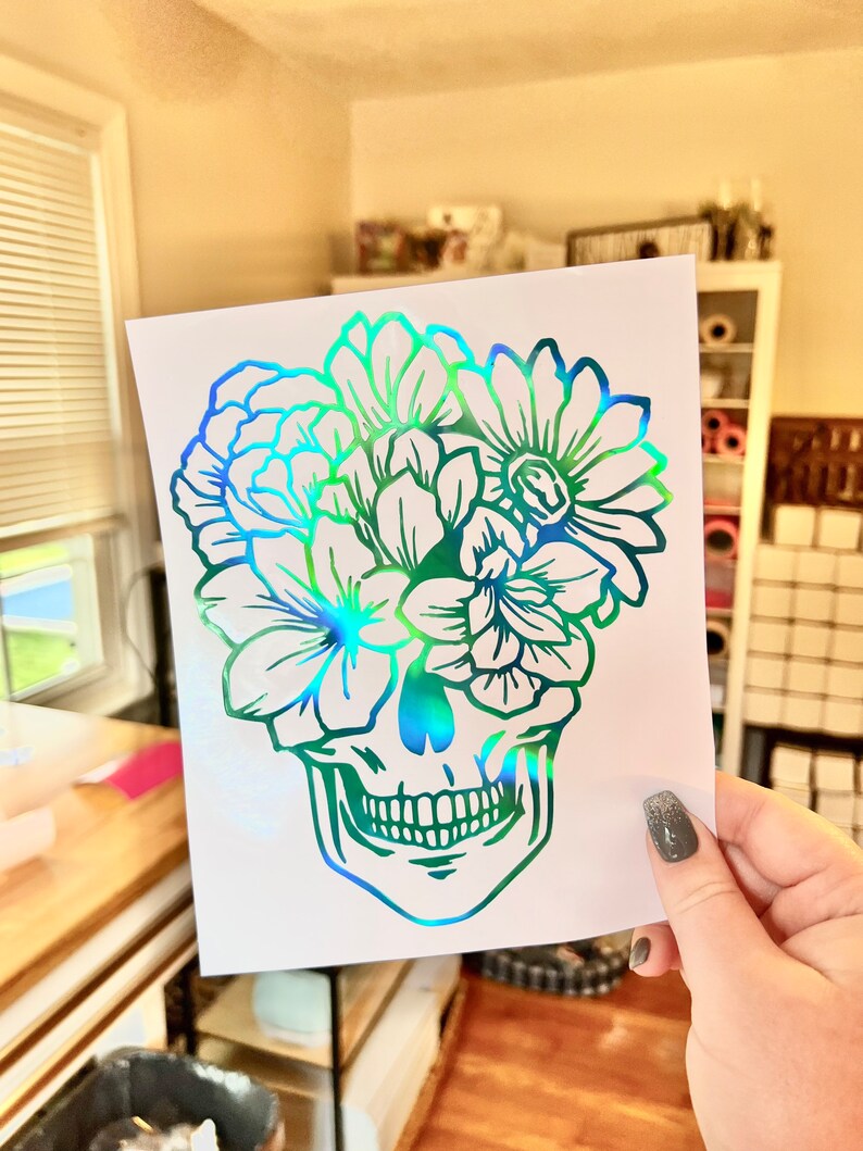 Puede incluir: Una pegatina hologr&aacute;fica con un dise&ntilde;o de calavera y elementos florales. La calavera est&aacute; delineada en verde y azul, con flores que florecen desde la parte superior. La pegatina se sostiene sobre un fondo brillante.
