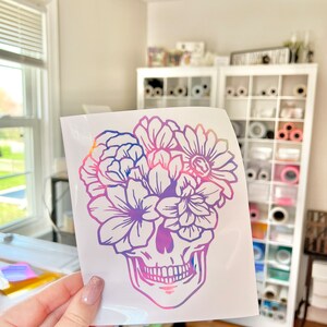 Puede incluir: Una calcoman&iacute;a de vinilo blanco con un dise&ntilde;o de calavera con una corona floral. La calavera y las flores est&aacute;n delineadas en un degradado de tonos rosas, morados y azules. La calcoman&iacute;a se sostiene frente a una habitaci&oacute;n luminosa.