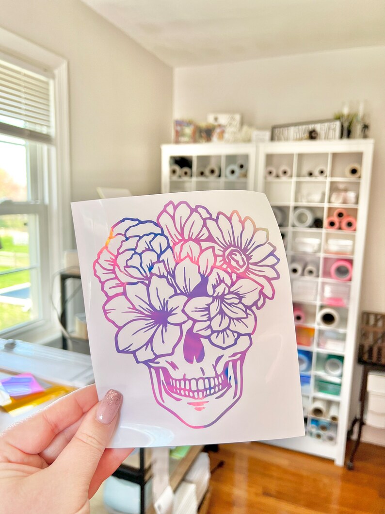 Puede incluir: Una calcoman&iacute;a de vinilo blanco con un dise&ntilde;o de calavera con una corona floral. La calavera y las flores est&aacute;n delineadas en un degradado de tonos rosas, morados y azules. La calcoman&iacute;a se sostiene frente a una habitaci&oacute;n luminosa.