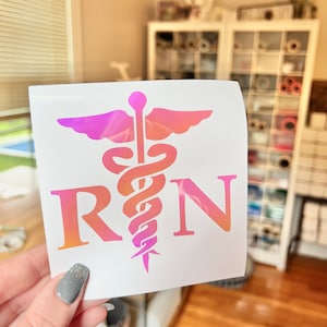 May include: A square decal featuring a pink and orange gradient caduceus symbol centered between the letters R and N. The design is printed on a white background, showcasing a medical theme with a smooth, colorful finish on the graphic elements.