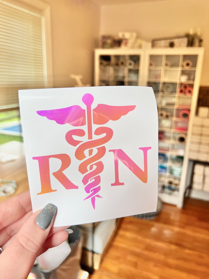 May include: A square decal featuring a pink and orange gradient caduceus symbol centered between the letters R and N. The design is printed on a white background, showcasing a medical theme with a smooth, colorful finish on the graphic elements.