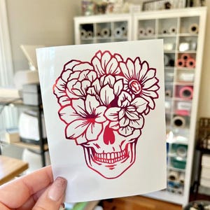 May include: A vinyl decal on a white backing sheet featuring a gradient red and pink skull design. The skull is adorned with a large arrangement of various flowers covering the upper portion, rendered in a bold, clean-lined graphic style.