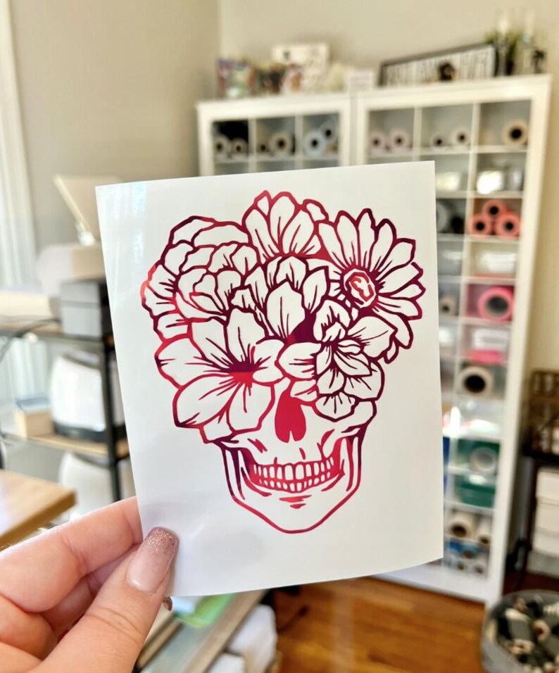 May include: A vinyl decal on a white backing sheet featuring a gradient red and pink skull design. The skull is adorned with a large arrangement of various flowers covering the upper portion, rendered in a bold, clean-lined graphic style.