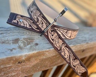 May include: A brown leather belt with a floral tooled design. The belt has a silver buckle and is shown on a wooden surface.