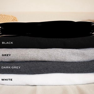 May include: A stack of four t-shirts in different colors: black, grey, dark grey, and white. The shirts are folded and stacked on top of each other.