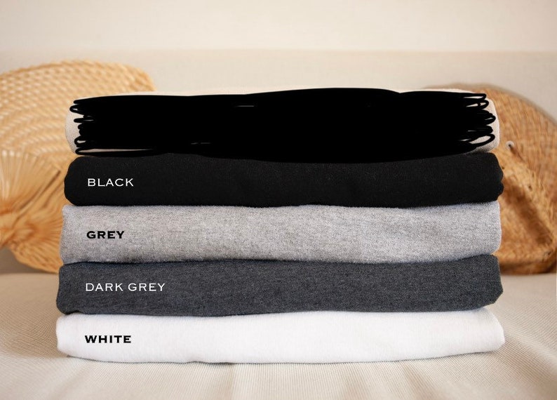 May include: A stack of four t-shirts in different colors: black, grey, dark grey, and white. The shirts are folded and stacked on top of each other.