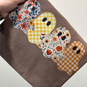 May include: Brown fabric with four ghost-shaped appliqués. The appliqués feature floral and gingham patterns in orange, yellow, and white. Each ghost has two black oval eyes. The fabric is held by a hand.