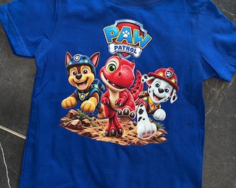 Puede incluir: Una camiseta azul real de manga corta con un estampado gráfico del logotipo de Paw Patrol sobre tres personajes: un perro marrón con uniforme azul, un dinosaurio rojo y un perro blanco con manchas negras y casco de bombero rojo.